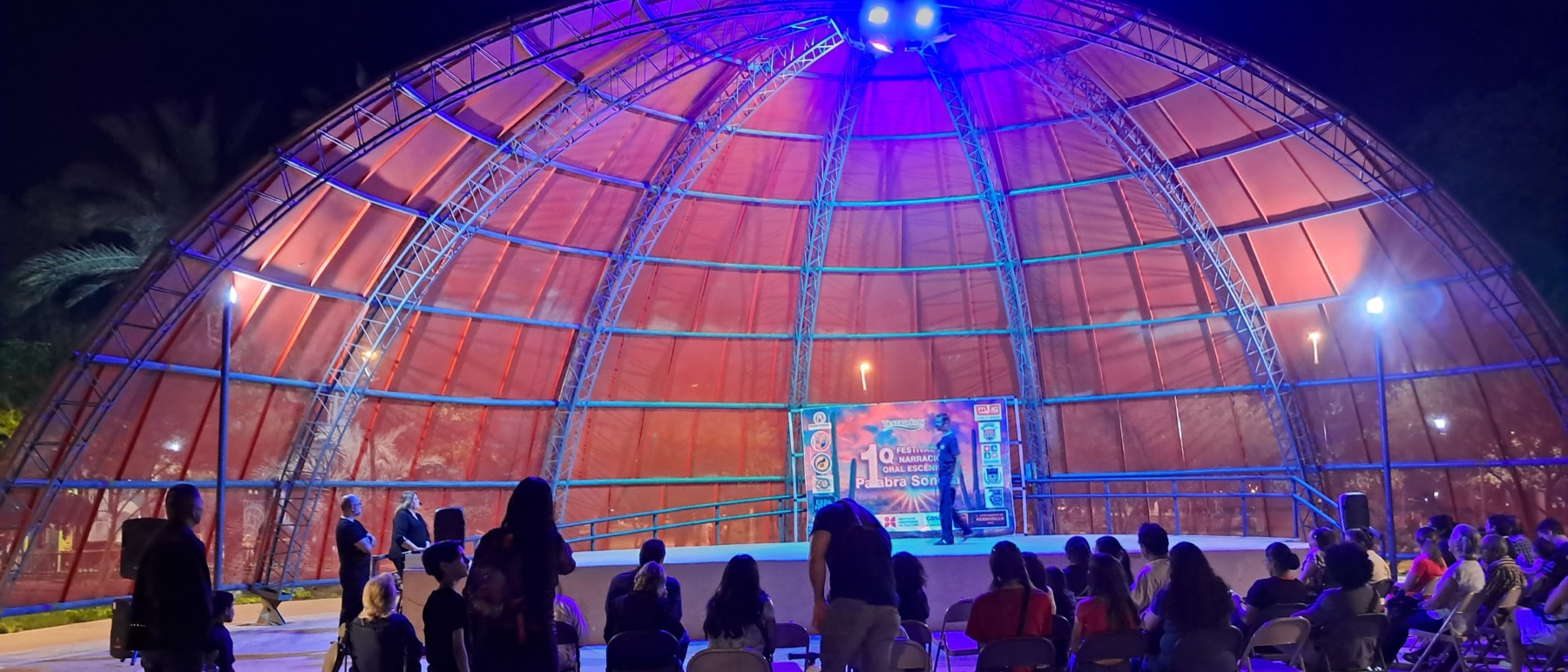 El domo del Parque Madero iluminado de morado y rojo por la noche, con el escenario del festival al fondo y el público sentado en silueta.