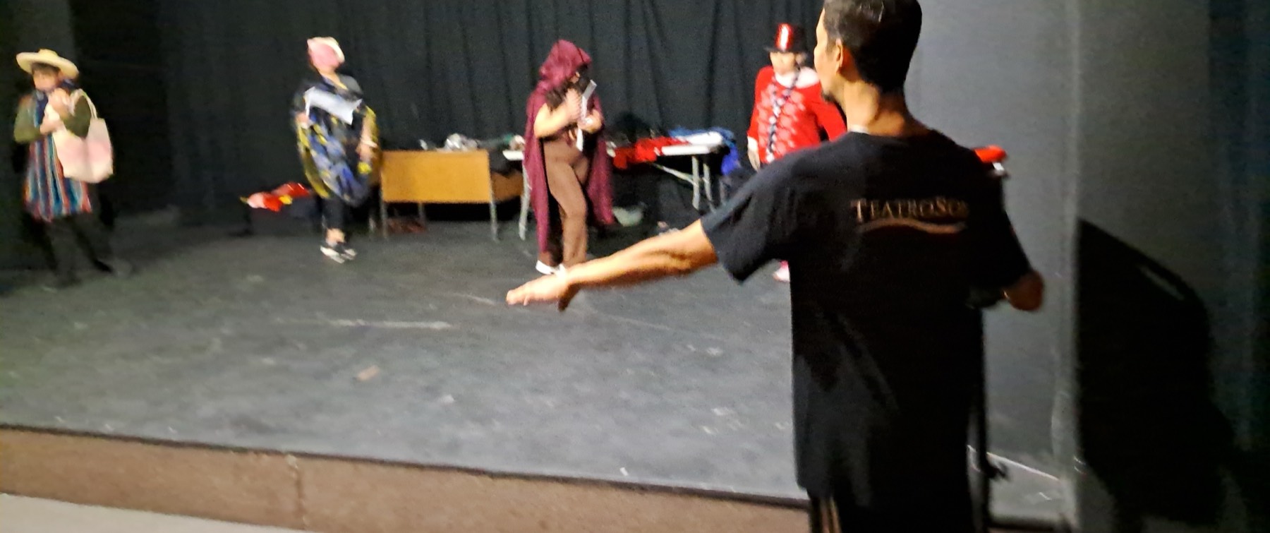 Escenario de cortinas negras durante un taller: participantes adultos en personaje al fondo y, de espaldas en primer plano, alguien dirigiendo la escena con playera de TeatroSon.