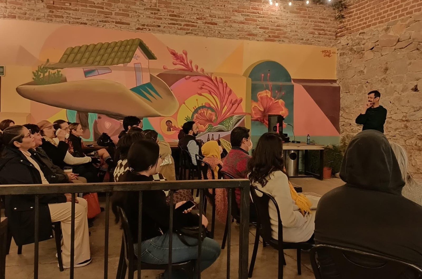 Julio Patricio narrando de pie ante público adulto sentado, con un mural de colores al fondo.