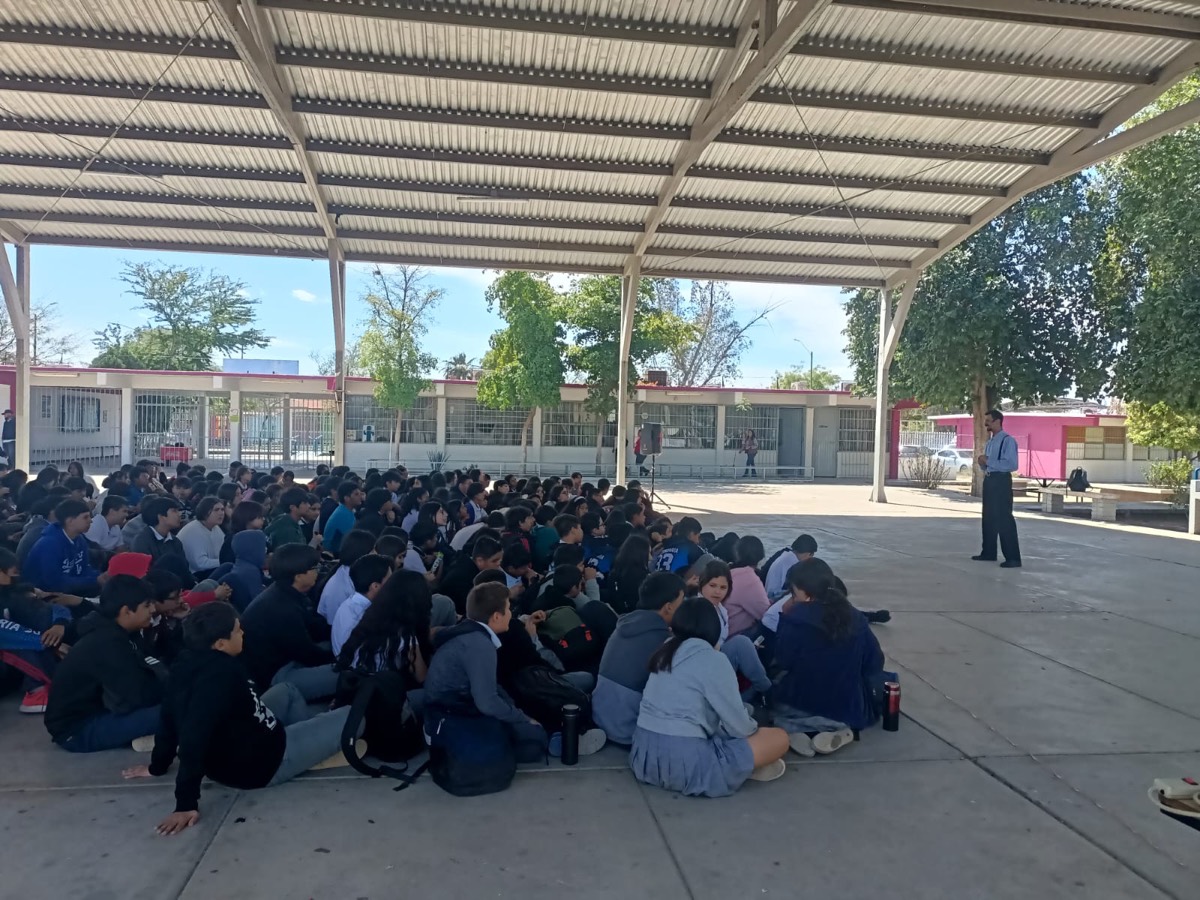 Vista general del patio techado de una secundaria: cientos de estudiantes sentados en el piso, de espaldas y a distancia, y Julio Patricio de pie al fondo.