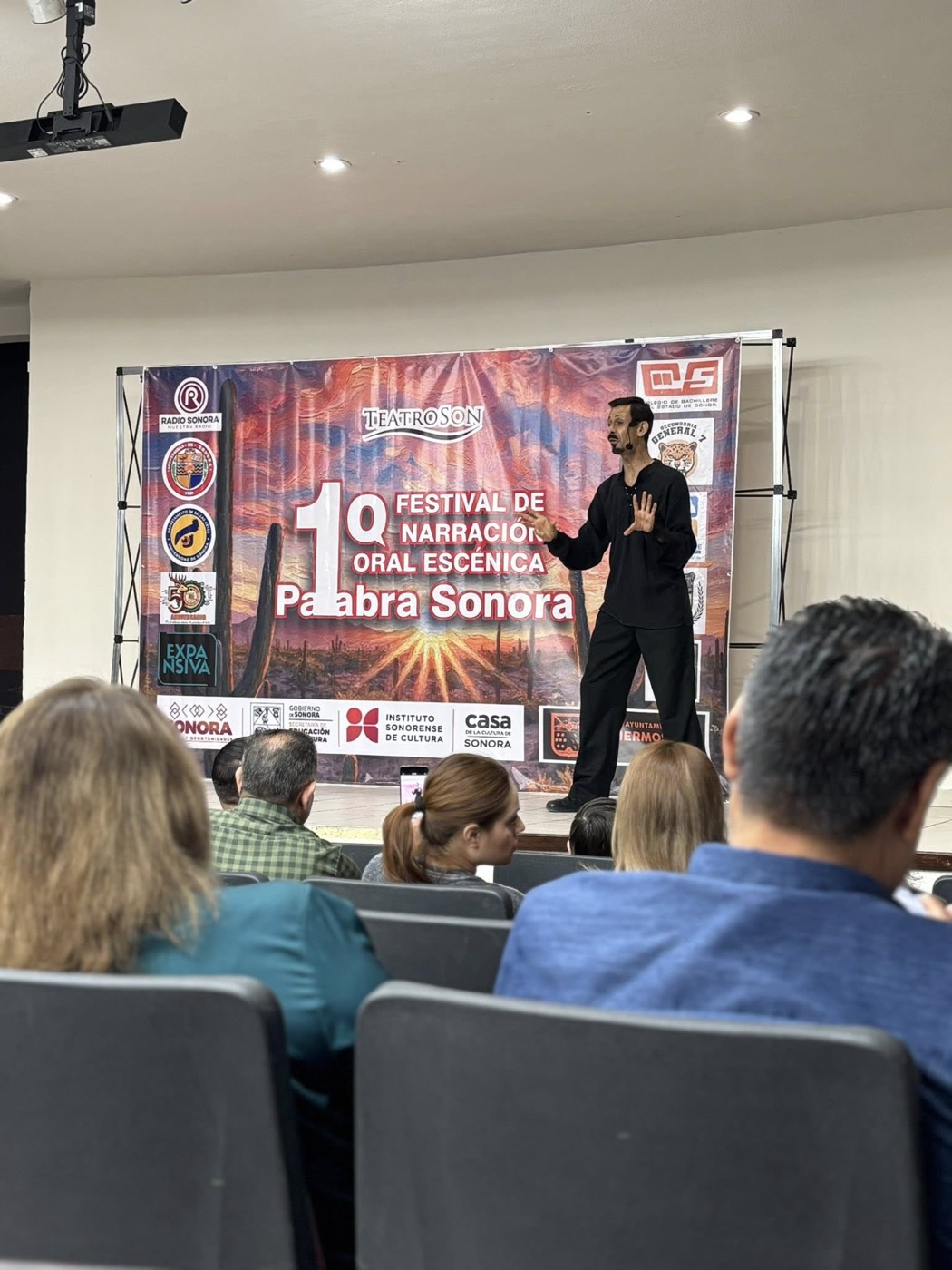 Julio Patricio narrando sobre el escenario en la función de gala, con el público de espaldas en primer plano.