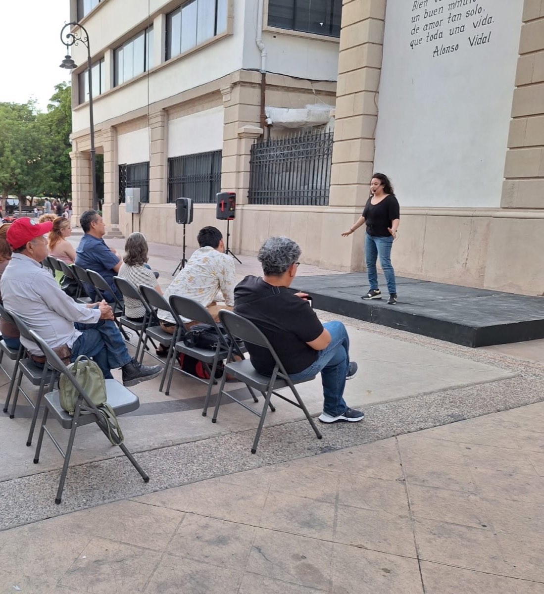 Una narradora de pie sobre la tarima en la calle, frente a público sentado en sillas plegables, junto a un muro con un poema de Alonso Vidal.
