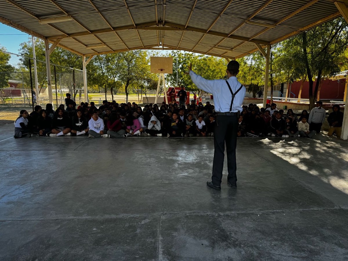 Julio Patricio de espaldas, con un brazo levantado, frente a un grupo grande de estudiantes sentados en el patio techado de una secundaria.